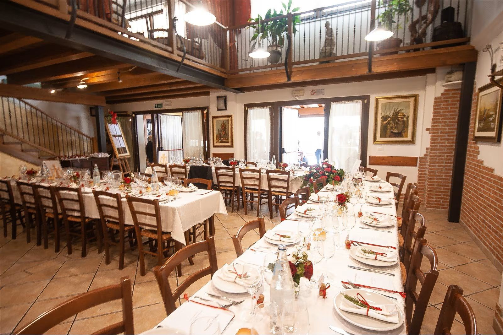 Sala da banchetto rustica con lunghi tavoli apparecchiati, sedie in legno, pareti in mattoni e calde luci sospese.