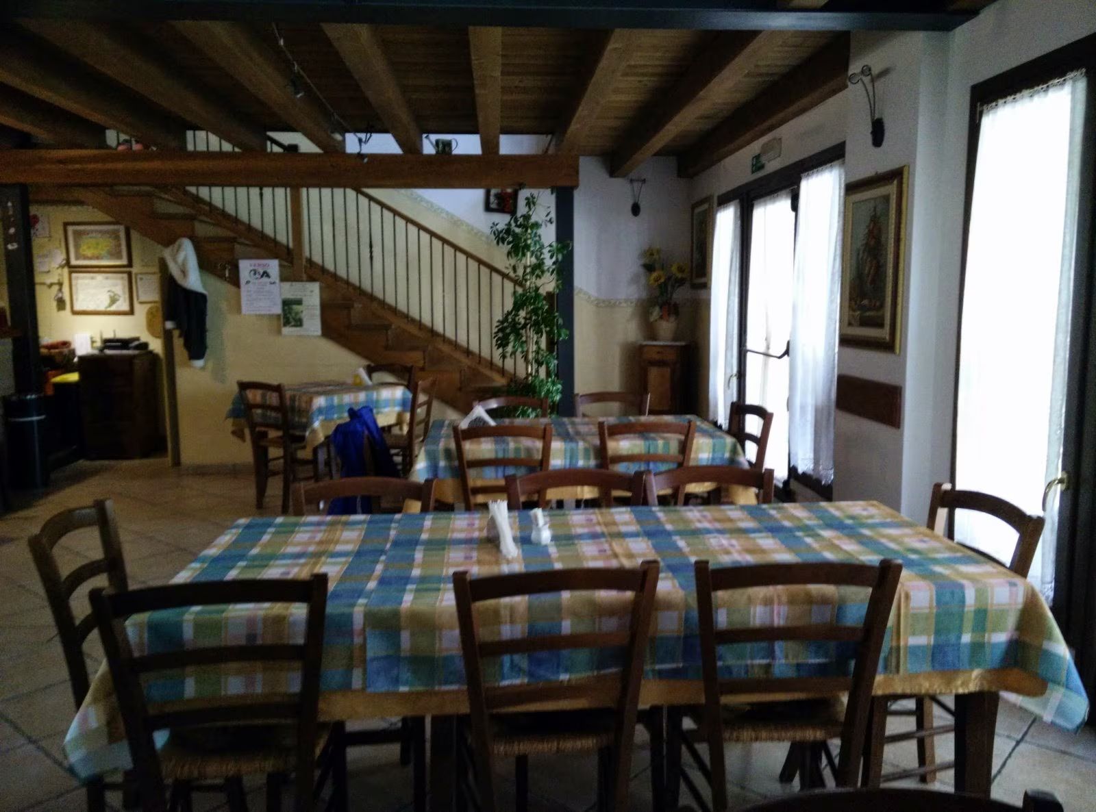 Sala da pranzo rustica con tovaglie a quadri, sedie in legno, scala e grandi finestre.