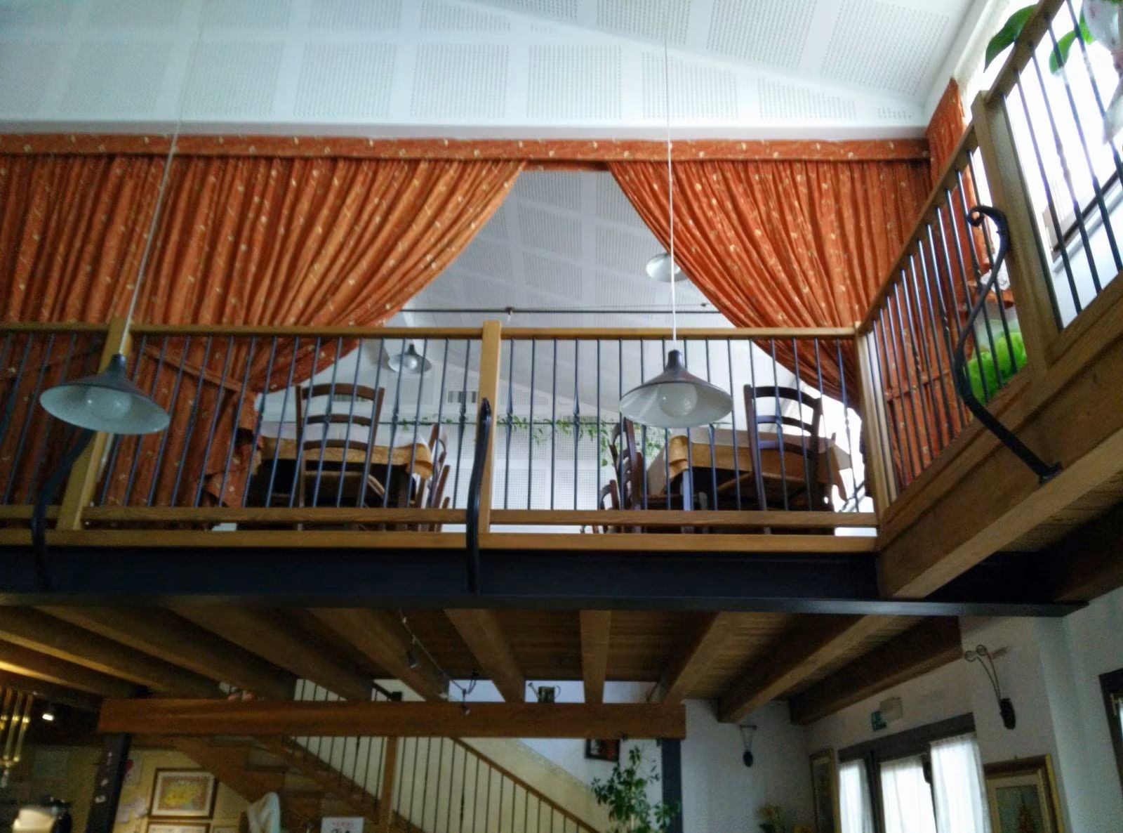 Zona pranzo in stile loft con soppalco in legno, tende arancioni e tavolini sotto la ringhiera.