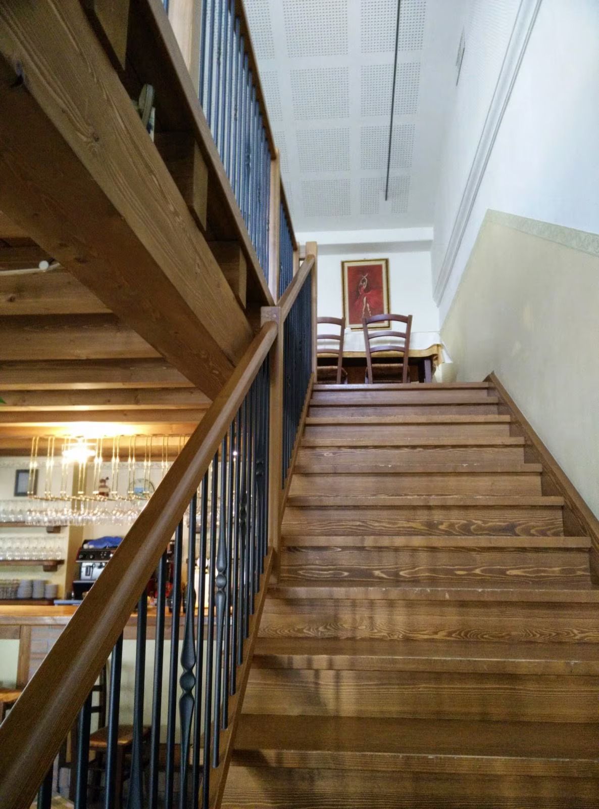 Scala in legno all'interno di un edificio, con sedie sul pianerottolo superiore e ringhiera in metallo.