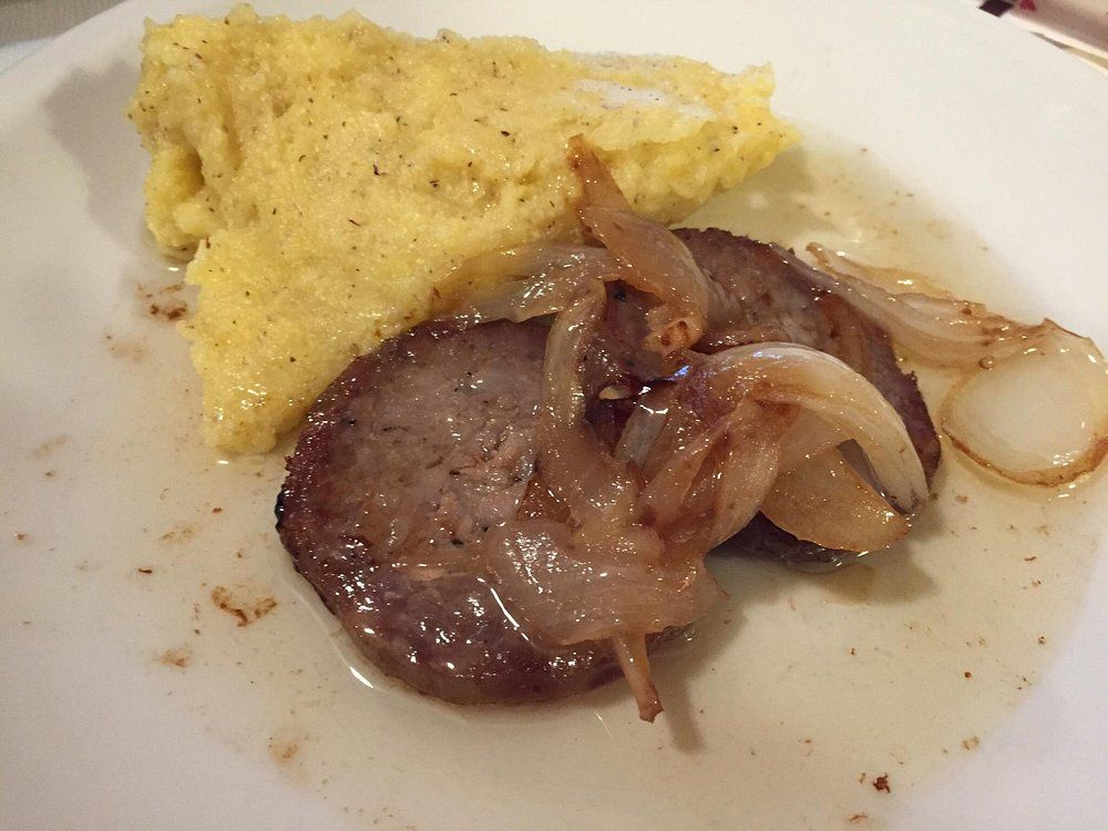 Piatto con una fetta di carne scottata, guarnita con cipolle e accompagnata da una salsa gialla simile alla polenta.