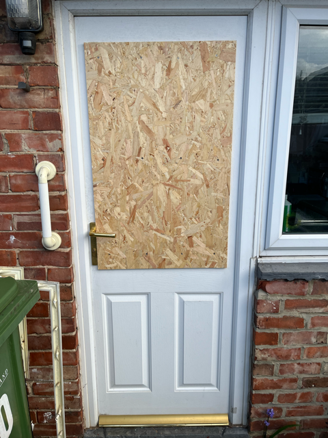 Front door boarded with plywood, white lower panel, next to brick wall and window.
