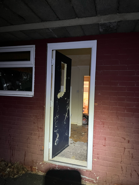 Damaged front door ajar on a red brick house, with light visible inside