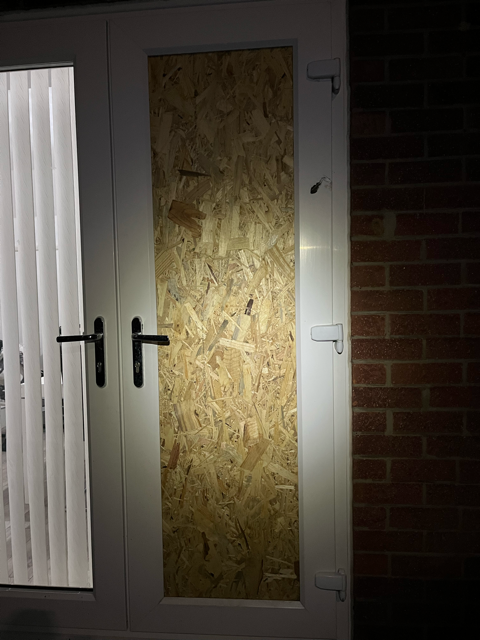 Boarded glass door beside a white shutter, with black handles and brick wall background