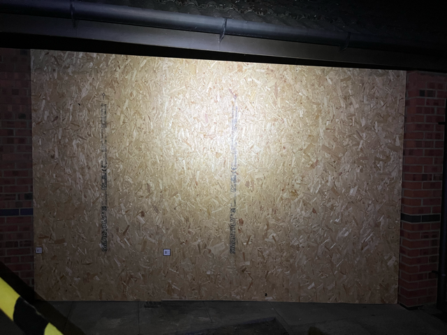 Plywood board covering a doorway or window in a dimly lit brick-walled area.