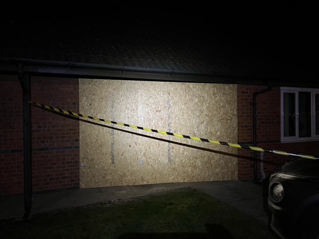 Caution tape stretched across a brick building at night, lit by a bright flashlight or spotlight.