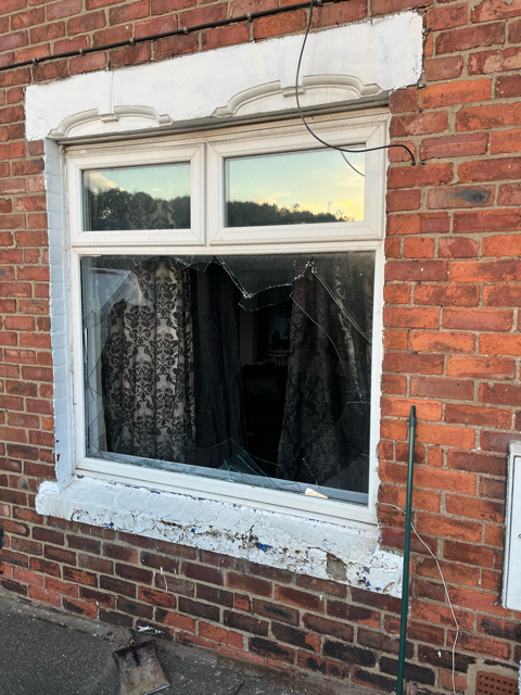 Broken white-framed window on a brick house with a cracked lower pane and closed curtains inside