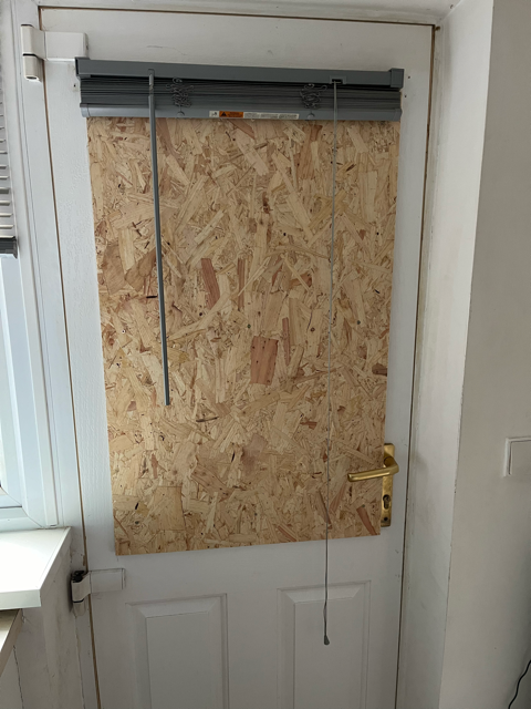 Boarded-up white door with OSB panel and a gray blind at the top