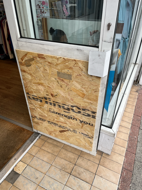 Plywood panel covering a damaged storefront window and door beside a tiled sidewalk
