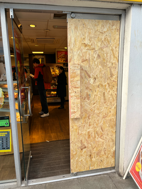 Store entrance with one glass door blocked by plywood; people visible inside.