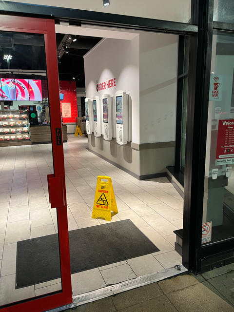 Store entrance with glass doors, white wall-mounted kiosks, and a yellow wet-floor caution sign on the floor.