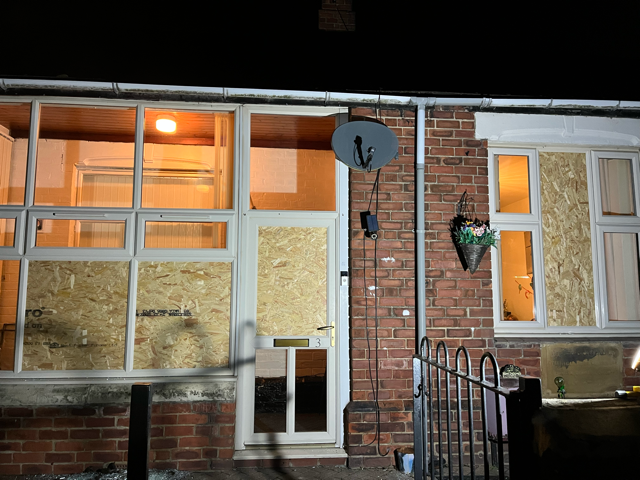 Brick house front at night with lit windows, boarded doors and windows, a satellite dish, and a small porch gate
