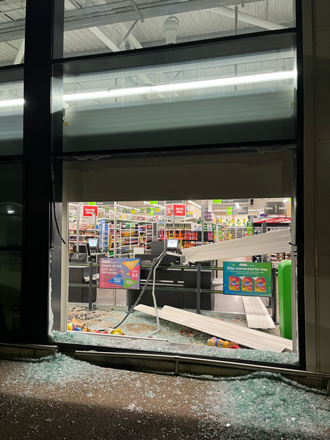 Broken storefront window with shattered glass and a damaged entrance at a brightly lit store.