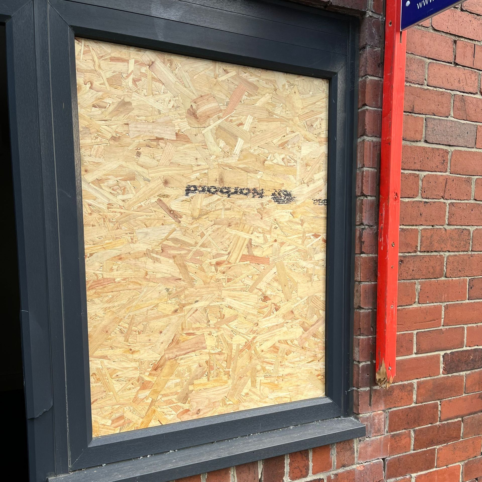 Window boarded up with particle board on a brick building with a dark gray frame.