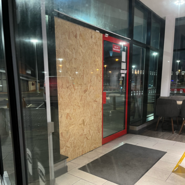 Boarded-up storefront with a red-framed glass door and dark entrance mat at night