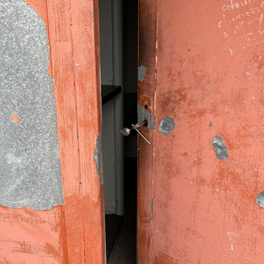 Red, weathered door slightly ajar, revealing a narrow interior space. The door has key in the lock.