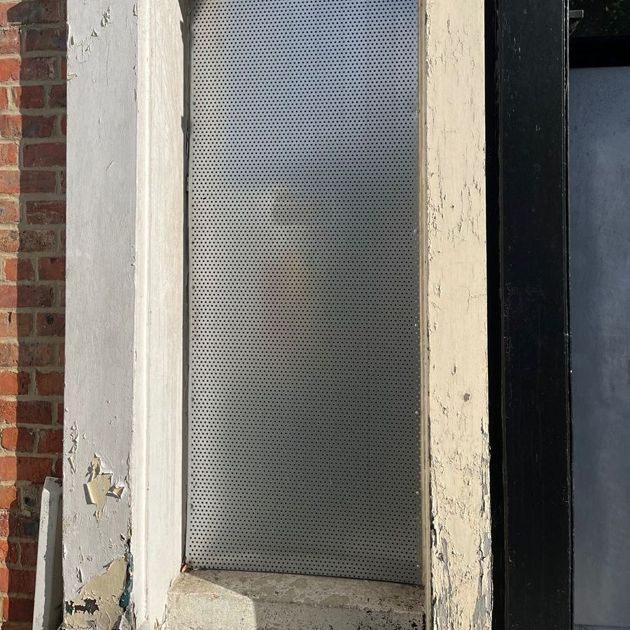 Perforated metal panel set into a weathered architectural recess, flanked by painted concrete and brick.