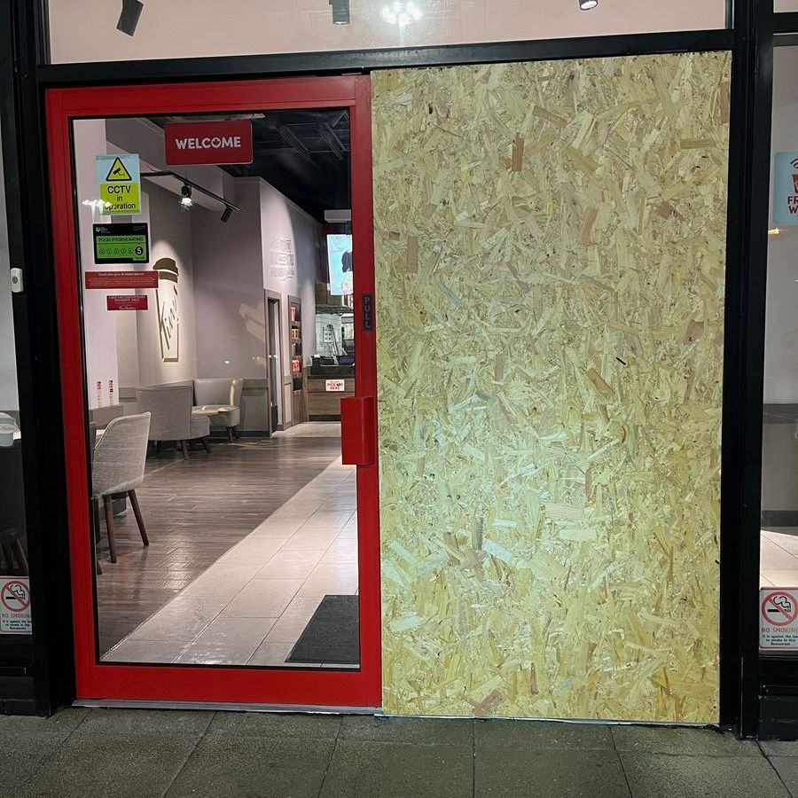 A partially boarded-up store entrance with a red door frame. A plywood panel covers the right side.
