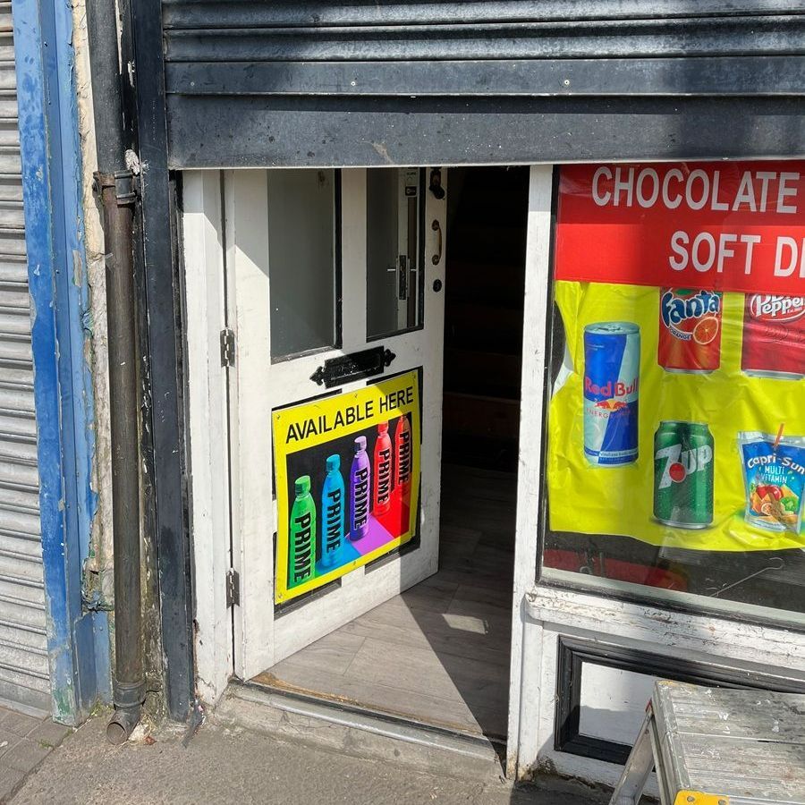 Shopfront with open white door, Prime drink advertising, soft drink poster. Shutter partially lowered.