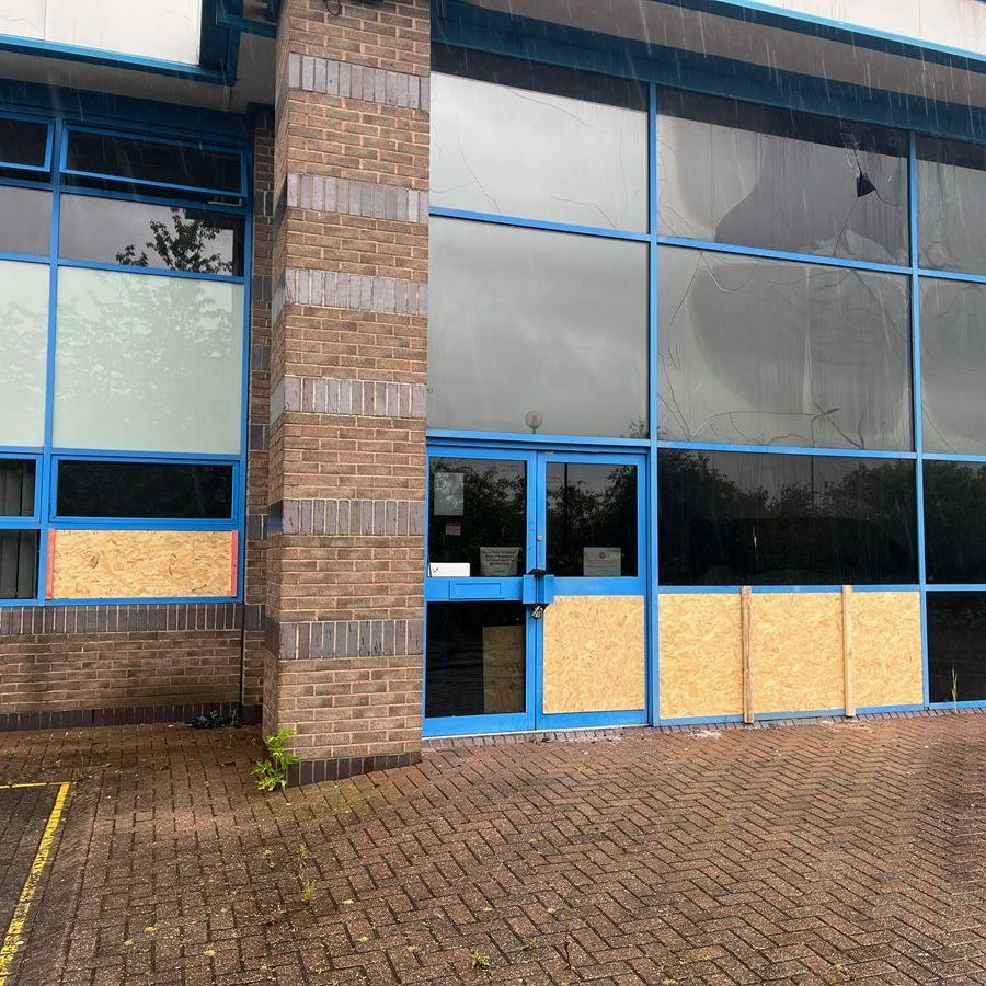 Building exterior with brick facade, large windows, and blue frames. Some windows boarded up.