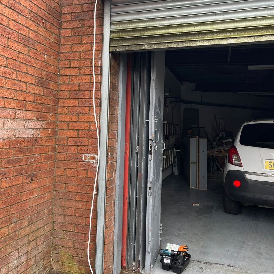 Partially open garage door. White car parked inside. Brick wall on left. Tools on the floor.