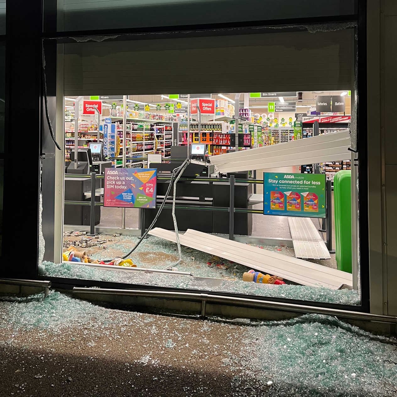 Shattered grocery store window with glass debris on the ground; inside, checkout counters and merchandise visible.