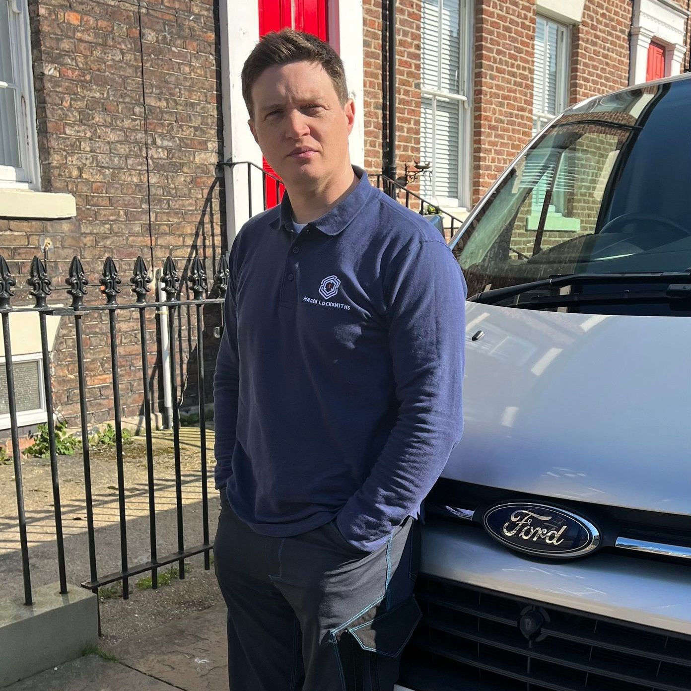 Man in blue shirt, standing next to a silver Ford van, on a street with a brick building and a wrought-iron fence.