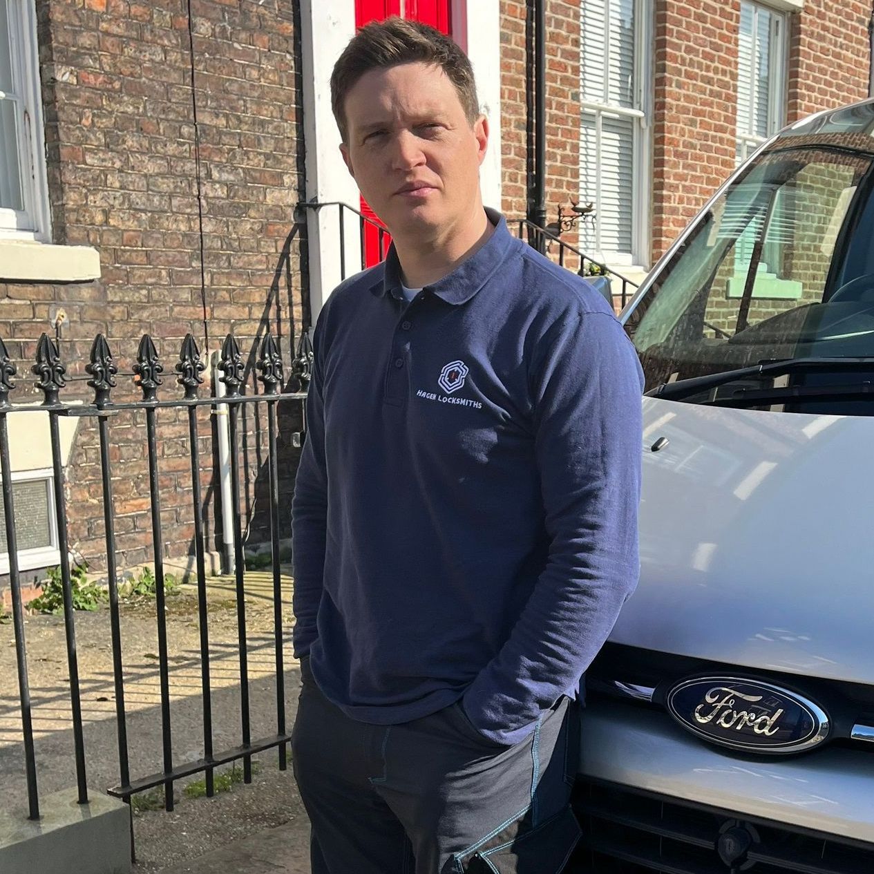 Man in blue shirt, hands in pockets, beside a white van parked in front of a brick building.