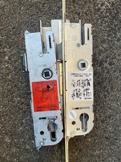 Two mortise door locks side by side, one silver and one brass, on a rough pavement surface.