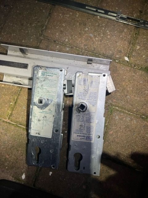 Two metal door lock mechanisms on a brick patio, one dark and one silver, lying side by side.
