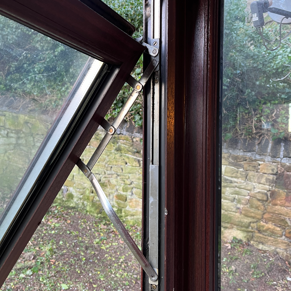 Brown casement window open, revealing metal hinges and a glimpse of an outdoor stone wall and greenery.