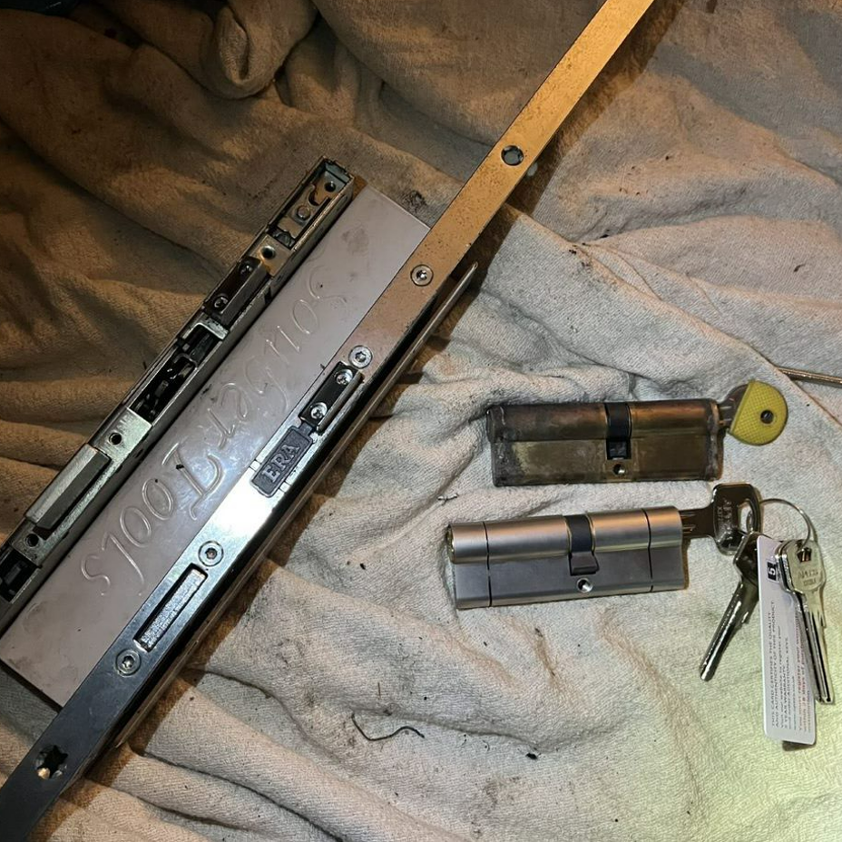 Lock hardware: door lock mechanism, two cylinders, keys, and a long metal bar, on a light cloth.