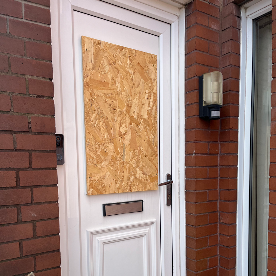 Door with plywood covering, brick building exterior. White door frame, motion sensor, and doorbell.