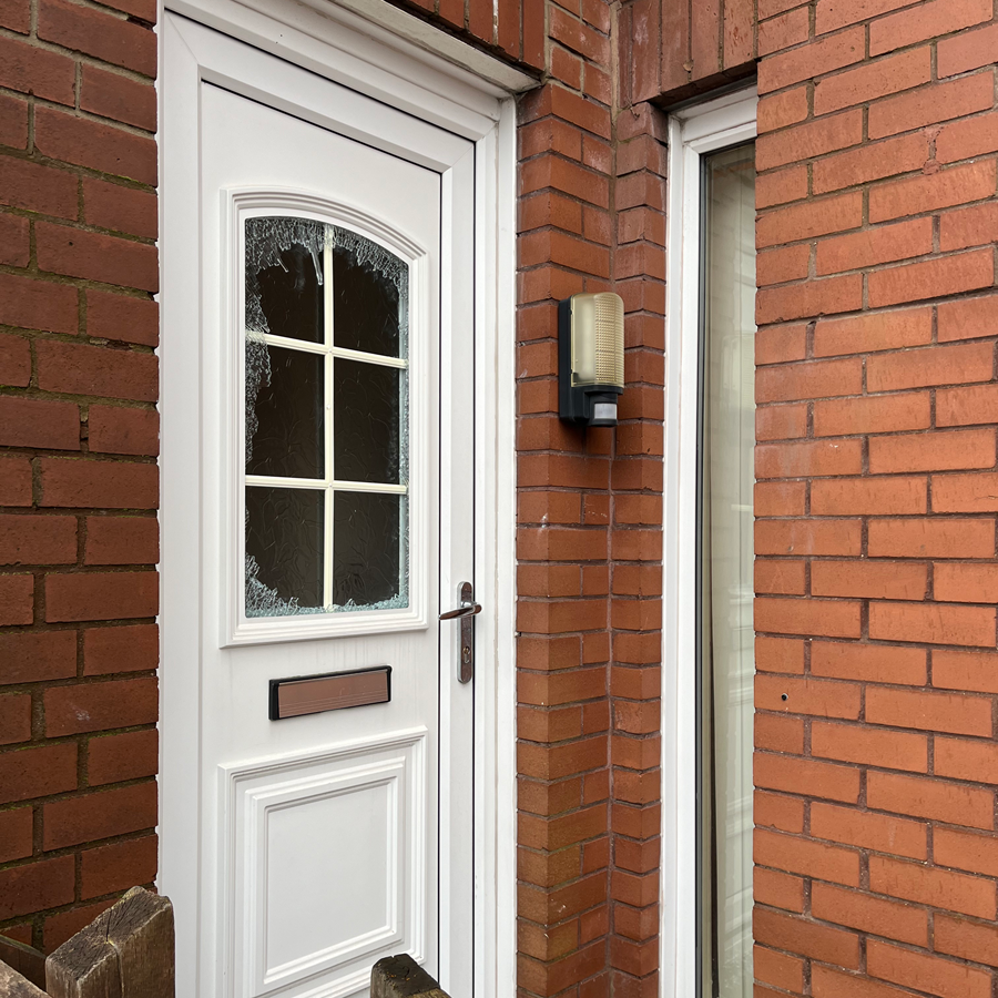 White front door with broken glass, set in a brick wall. A light is mounted on the wall.