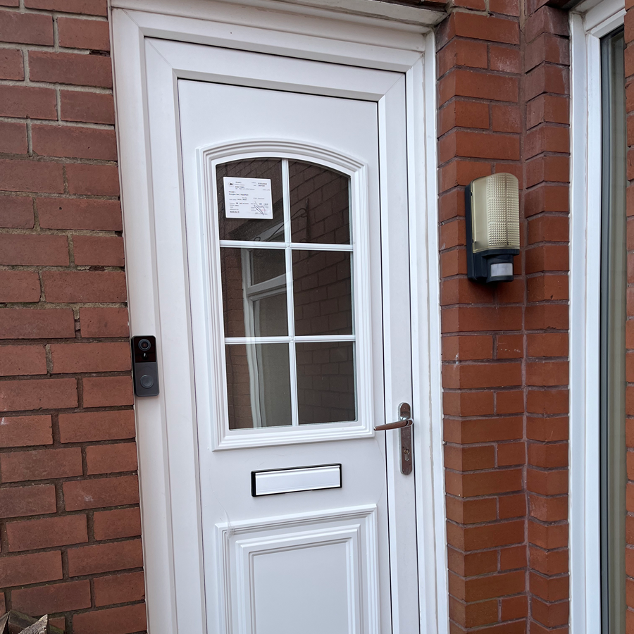 White front door with window panels, mailbox, doorbell, and outdoor light on a brick wall.