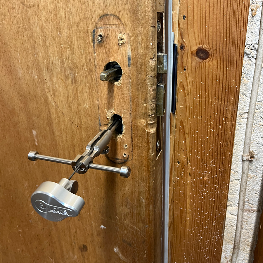 Door with lock removed, showing a tool in the lock hole. Wood door is light brown with metal components.