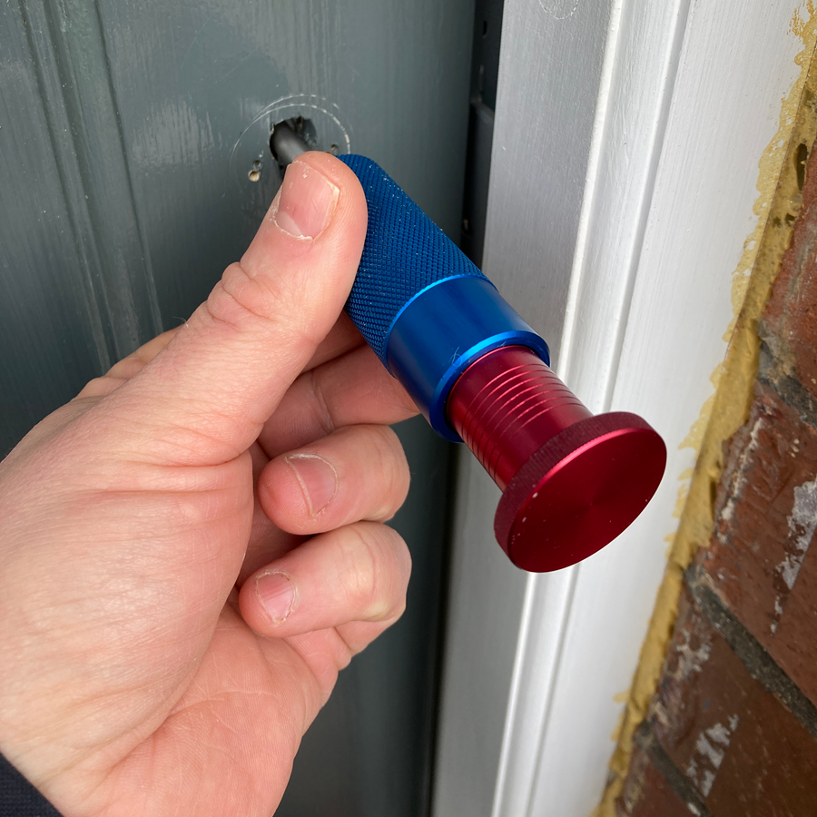 Hand holding a blue and red door lock pick tool inserted into a gray door.