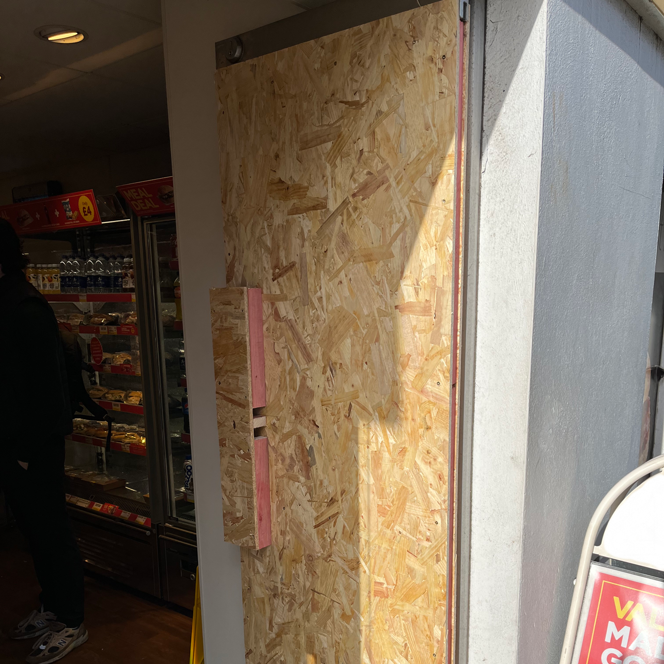 Boarded-up shop entrance. A plywood panel covers the door frame next to a white pillar.