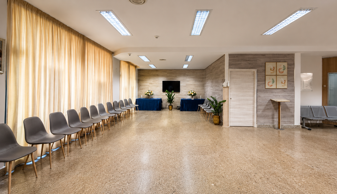 Sala d'attesa vuota con file di sedie, pareti beige, luci intense sul soffitto e piante in vaso in fondo.