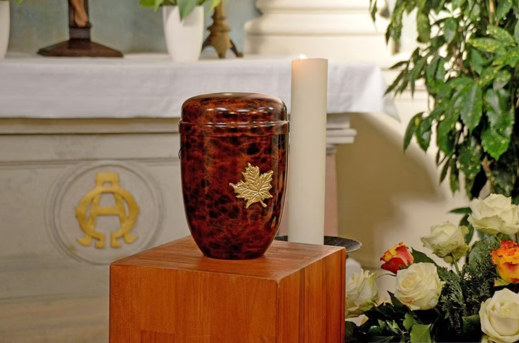 Una candela di vetro rossa accesa accanto a una candela a colonna bianca e fiori su un piedistallo di legno.