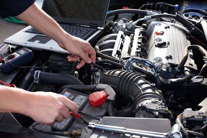 Meccanico al lavoro sotto il cofano aperto di un'auto, con attrezzi vicino al motore e alla batteria.