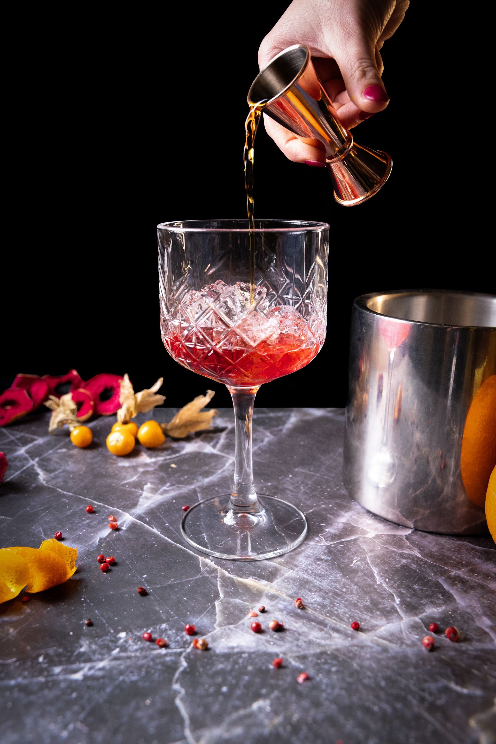 Un cocktail versato in un bicchiere di cristallo con ghiaccio. Una mano regge un misurino. Sfondo nero, con frutta e shaker nelle vicinanze.