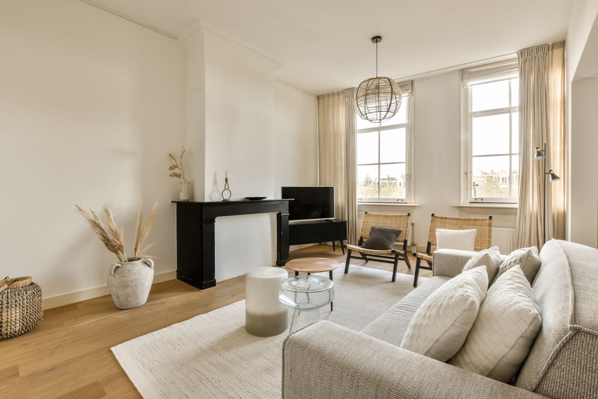 Bright living room with beige sofa, black fireplace, wooden chairs, and tall windows with curtains.