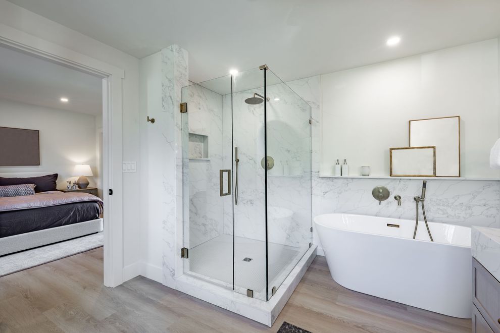Bright modern bathroom with glass shower, freestanding tub, and adjoining bedroom visible through open doorway