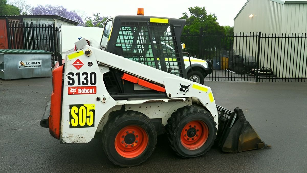 White skid-steer loader with orange wheels parked on wet pavement beside a fence and warehouse.