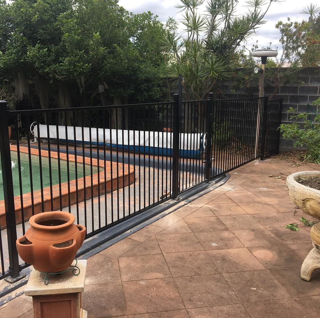 Backyard with a blue swimming pool, black fence, lounge chair, and tall hedges under a clear sky