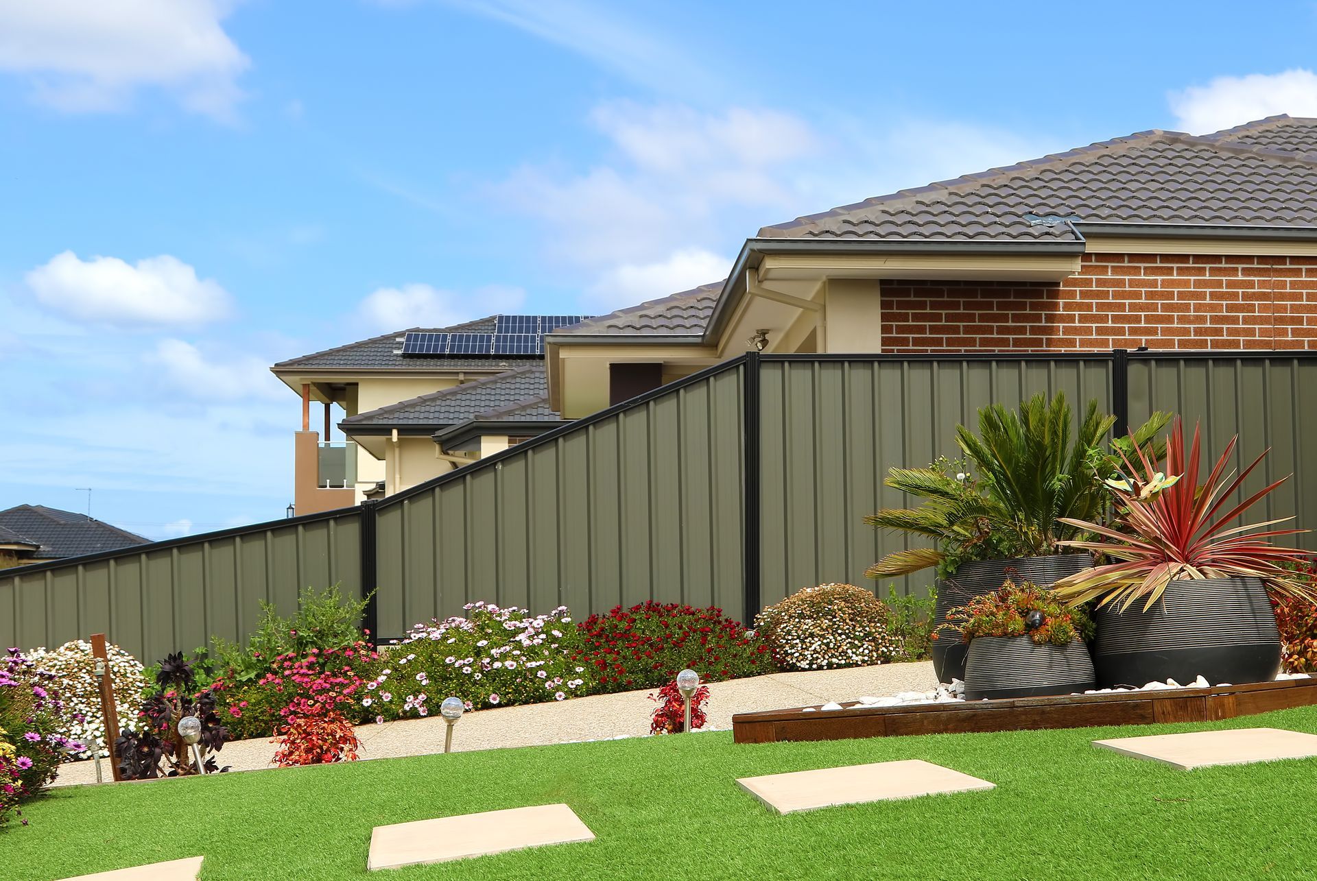 Modern suburban houses behind a green fence, with a landscaped front yard and blue sky.