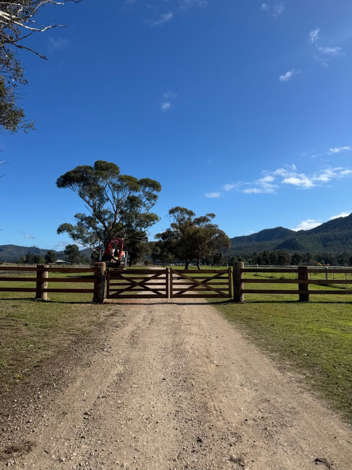 Dirt driveway leading to a wooden gate in a sunny rural field with hills and a tree under a blue sky