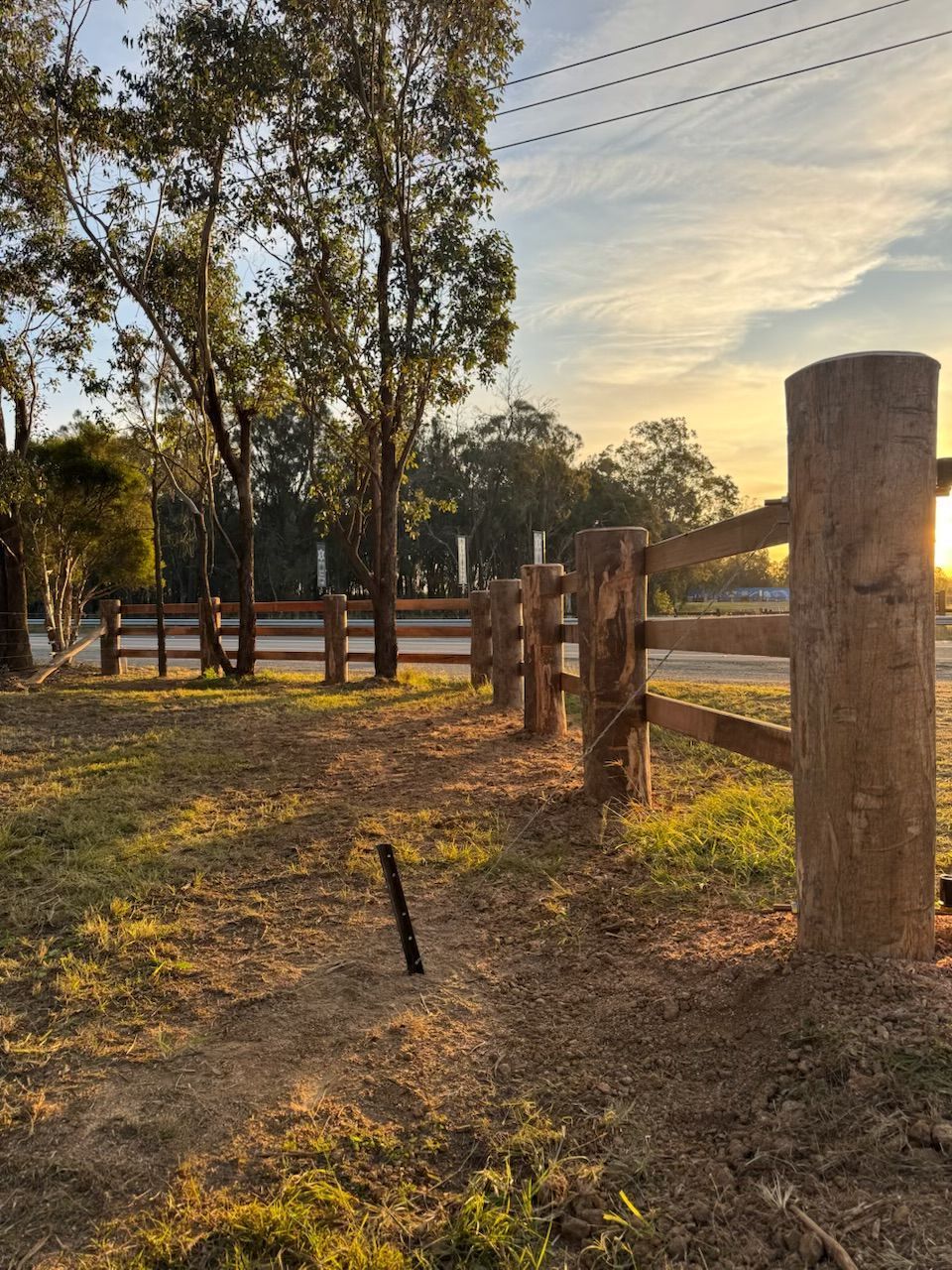 Wooden fence posts along a sunlit park path at sunset, with trees and open field in the background.