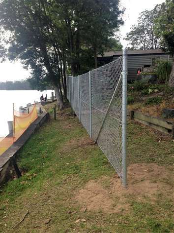 Chain-link fence along a grassy lakeside slope with trees and a dock on the left